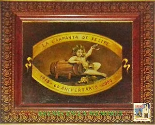 Azulejo enmarcado en madera con un querubín sentado sobre un barril y la leyenda: La Carpanta de Felipe, 1940, 65 aniversario, 2005.