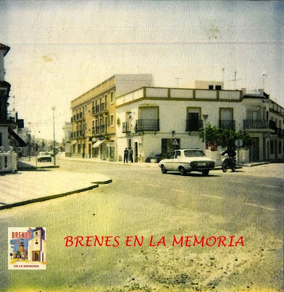 Fotografía antigua coloreada de la calle: casas encaladas de dos plantas haciendo esquina y coches de los años setenta aparcados.